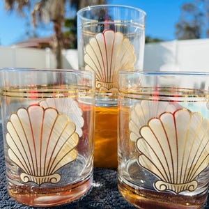 May include: Three clear drinking glasses with a gold rim and a gold seashell design. The glasses are arranged on a dark surface, with a bright blue sky in the background. The seashell design is in the center of each glass.