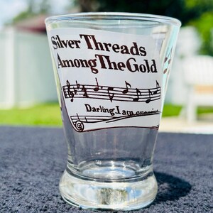 May include: A clear glass shot glass featuring a black printed graphic of musical notes on a staff. The text reads Silver Threads Among The Gold and Darling I am growing. The glass has a slightly flared rim and a thick, weighted base design.