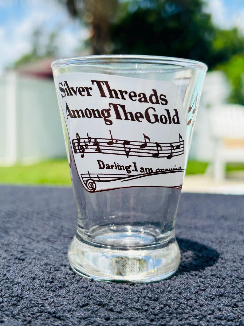 May include: A clear glass shot glass featuring a black printed graphic of musical notes on a staff. The text reads Silver Threads Among The Gold and Darling I am growing. The glass has a slightly flared rim and a thick, weighted base design.