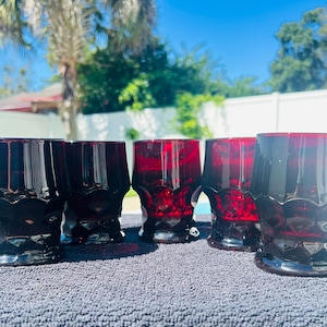 May include: A set of five deep red glass tumblers featuring a faceted, geometric pattern around the base and a smooth, cylindrical upper section. The glasses are made of translucent ruby-colored glass that catches the light, showing a distinct texture.