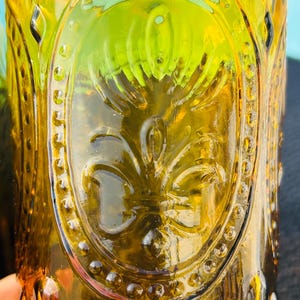 May include: Close-up of a vintage amber-colored glass. The glass features an embossed design of a fleur-de-lis within an oval frame, surrounded by small, raised dots. The glass has a textured surface and a slight green hue.