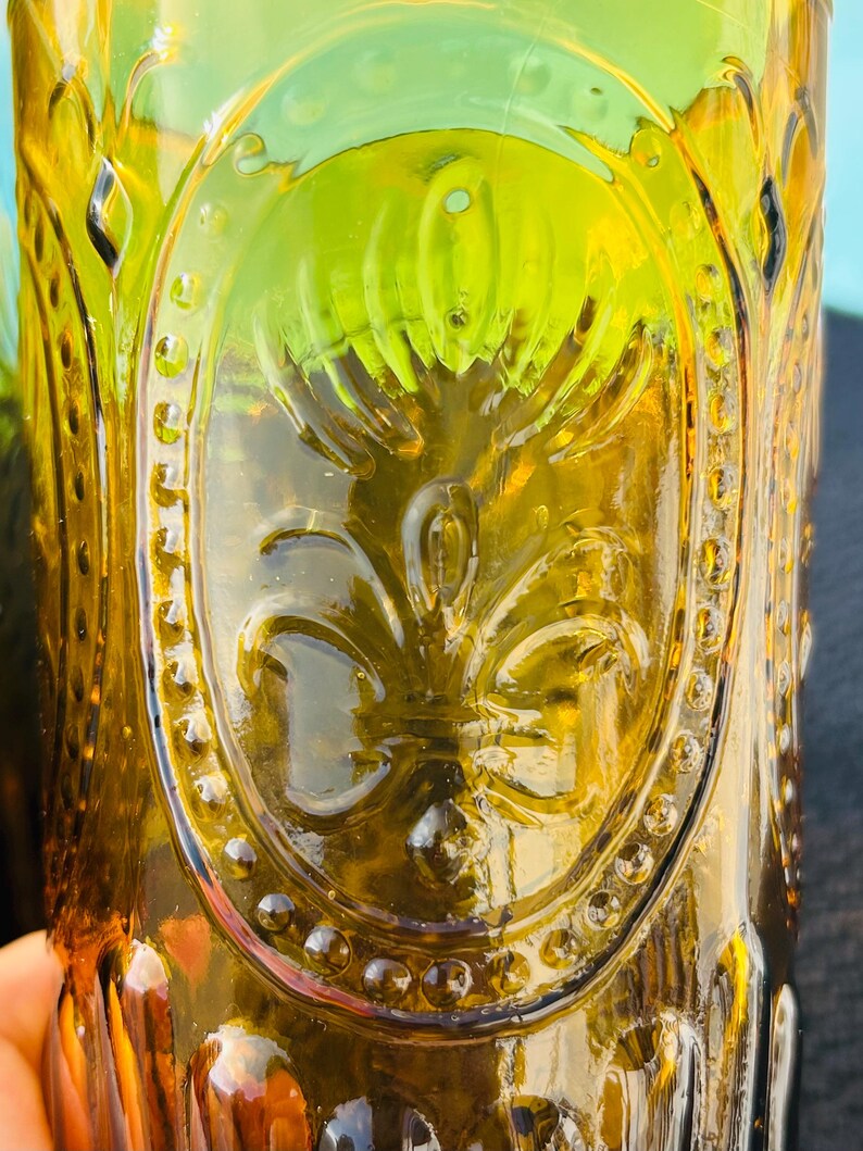 May include: Close-up of a vintage amber-colored glass. The glass features an embossed design of a fleur-de-lis within an oval frame, surrounded by small, raised dots. The glass has a textured surface and a slight green hue.