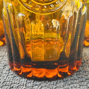 May include: Close-up of amber-colored drinking glasses. The glasses have a textured, ribbed design and a decorative pattern near the top. The glass is thick and has a rich, warm color, with a darker amber hue at the base.