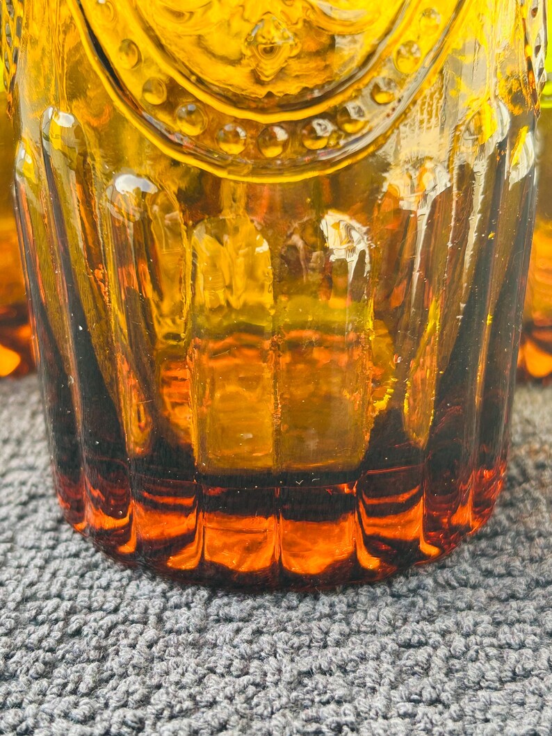 May include: Close-up of amber-colored drinking glasses. The glasses have a textured, ribbed design and a decorative pattern near the top. The glass is thick and has a rich, warm color, with a darker amber hue at the base.