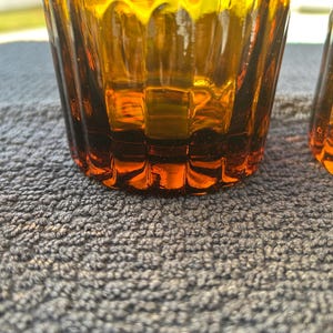 May include: Close-up of two amber-colored drinking glasses. The glasses have a textured, ribbed design and a slightly flared shape. The color transitions from a darker amber at the base to a lighter, golden hue towards the top. The glasses are on a gray surface.