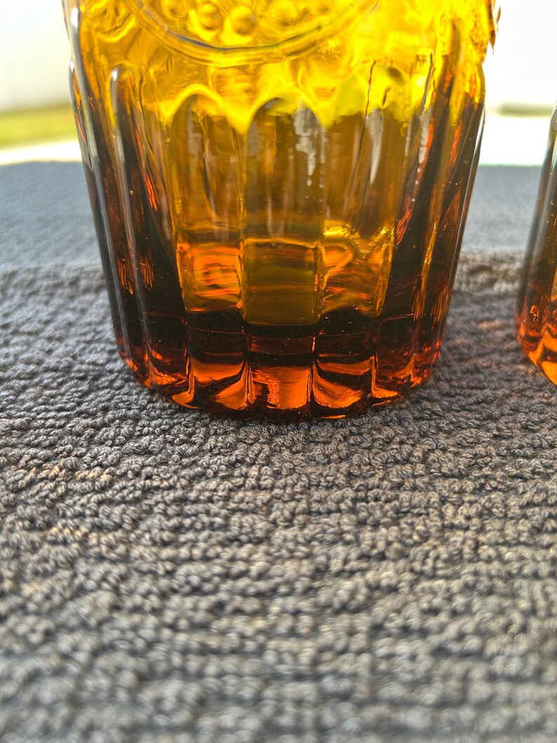 May include: Close-up of two amber-colored drinking glasses. The glasses have a textured, ribbed design and a slightly flared shape. The color transitions from a darker amber at the base to a lighter, golden hue towards the top. The glasses are on a gray surface.