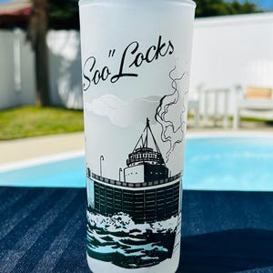 May include: A frosted glass tumbler with a black and white illustration of a ship and the text "Soo Locks". The design includes waves and a detailed depiction of the ship's structure. The glass is cylindrical.