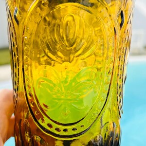May include: A vintage amber-colored glass with an embossed floral design. The glass has a tall, cylindrical shape and is partially filled with a green liquid. The glass has a decorative oval design in the center.
