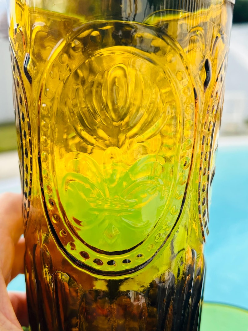 May include: A vintage amber-colored glass with an embossed floral design. The glass has a tall, cylindrical shape and is partially filled with a green liquid. The glass has a decorative oval design in the center.