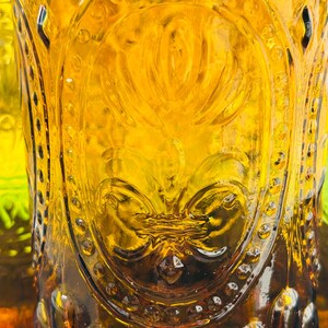 May include: Close-up of a vintage amber-colored glass with ornate embossed detailing. The glass features a central floral design and a repeating pattern of small circles around the rim. The glass has a textured surface and is partially filled.