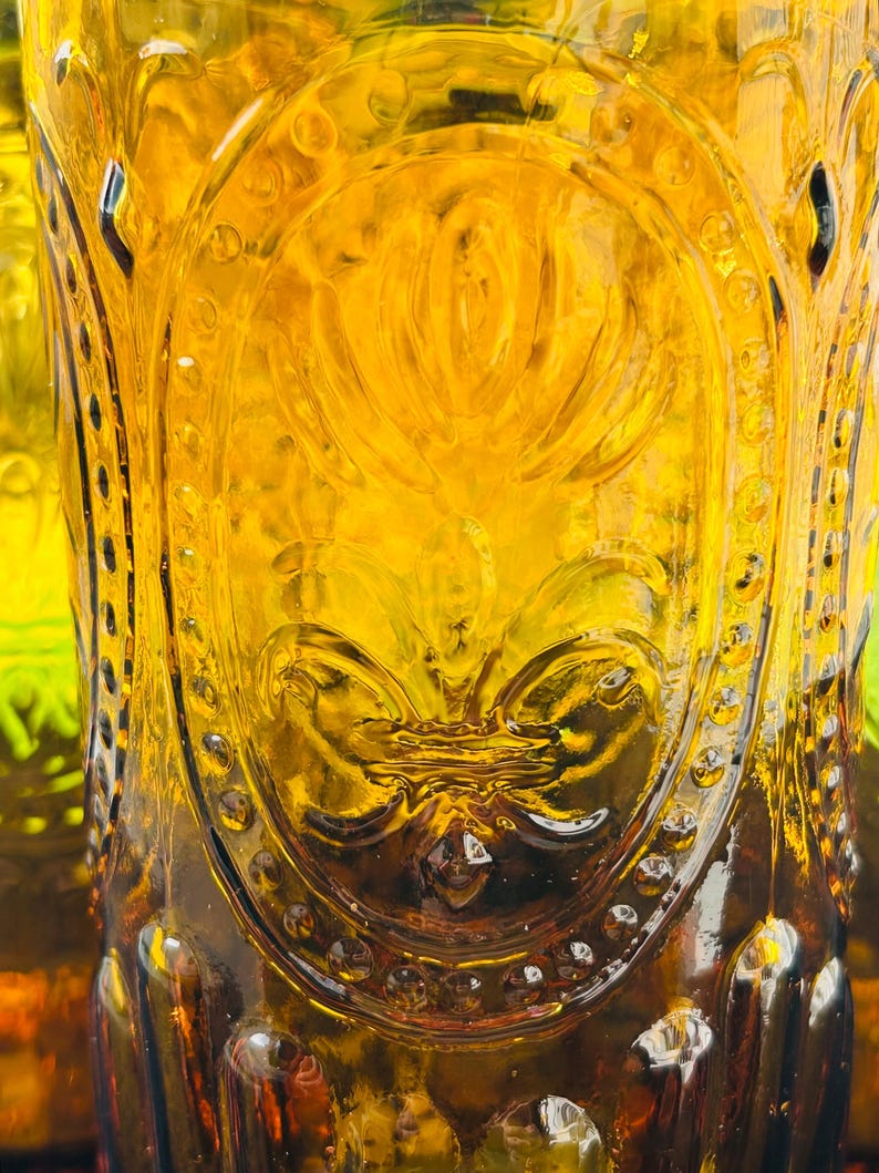 May include: Close-up of a vintage amber-colored glass with ornate embossed detailing. The glass features a central floral design and a repeating pattern of small circles around the rim. The glass has a textured surface and is partially filled.