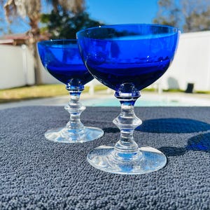 May include: Two cobalt blue stemmed glasses with a clear, ornate stem and base. The glasses are set on a dark blue textured surface, with a bright blue sky in the background. The glasses are empty.
