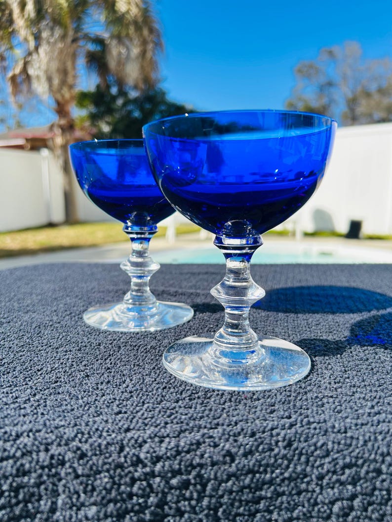 May include: Two cobalt blue stemmed glasses with a clear, ornate stem and base. The glasses are set on a dark blue textured surface, with a bright blue sky in the background. The glasses are empty.