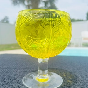 May include: A vibrant yellow glass goblet featuring a raised, textured relief pattern across the bowl. The stem and circular base are made of clear, smooth glass, providing a contrast to the bright, translucent yellow upper section of the vessel.