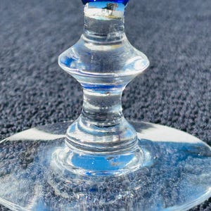 May include: Close-up of a clear glass wine goblet with a blue and white rim. The stem has a unique hourglass shape. The base is round and clear. The glass reflects the surrounding environment.