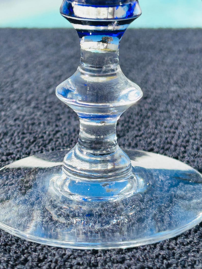May include: Close-up of a clear glass wine goblet with a blue and white rim. The stem has a unique hourglass shape. The base is round and clear. The glass reflects the surrounding environment.