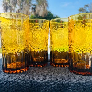 May include: Four amber-colored drinking glasses with embossed floral and geometric designs. The glasses are cylindrical with a slightly flared base. The glasses are sitting on a dark blue surface, with a bright blue sky and palm trees in the background.