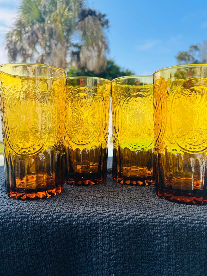 May include: Four amber-colored drinking glasses with embossed floral and geometric designs. The glasses are cylindrical with a slightly flared base. The glasses are sitting on a dark blue surface, with a bright blue sky and palm trees in the background.