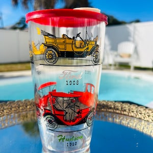 May include: Clear plastic tumbler with a red lid featuring vintage car illustrations. The tumbler has a yellow Ford car from 1908 and a red Hudson car from 1910. The tumbler is on a silver tray.