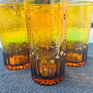 May include: Three amber-colored drinking glasses with embossed floral designs. The glasses have a tall, cylindrical shape with a decorative pattern and a textured base. The glasses are made of a transparent material, allowing light to pass through.