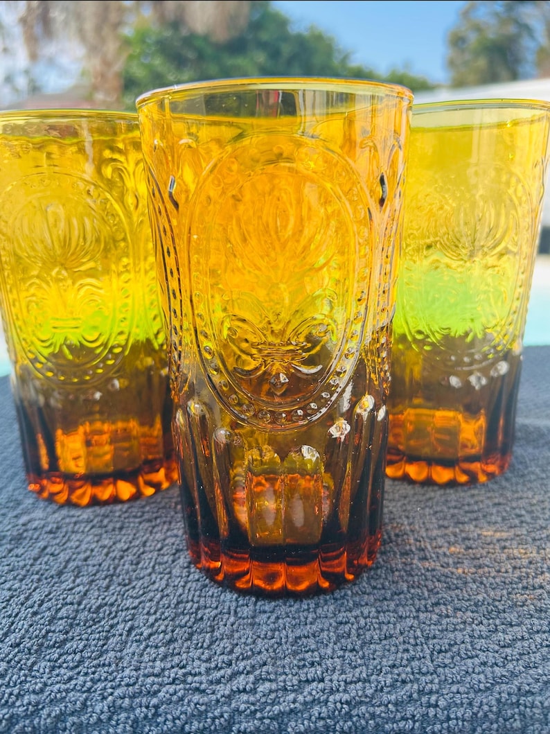 May include: Three amber-colored drinking glasses with embossed floral designs. The glasses have a tall, cylindrical shape with a decorative pattern and a textured base. The glasses are made of a transparent material, allowing light to pass through.