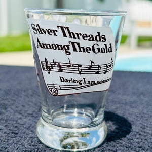 Könnte beinhalten: Ein klares Glas mit einem weißen Aufkleber, der den schwarzen Text Silver Threads Among The Gold sowie ein Notensystem mit Noten und den zusätzlichen Text Darling I am growing old in einer klaren, gut lesbaren Schriftart zeigt.