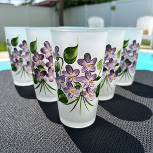 May include: Set of six frosted glass tumblers with hand-painted purple and green floral designs. The glasses are white with a frosted finish and feature a delicate floral pattern.