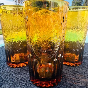 May include: Three amber-colored drinking glasses with a decorative embossed design. The glasses have a tall, cylindrical shape and a flared rim. The embossed design features a floral or fleur-de-lis pattern. The glasses are made of a transparent material, allowing light to pass through.
