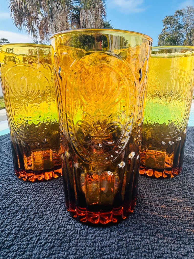May include: Three amber-colored drinking glasses with a decorative embossed design. The glasses have a tall, cylindrical shape and a flared rim. The embossed design features a floral or fleur-de-lis pattern. The glasses are made of a transparent material, allowing light to pass through.