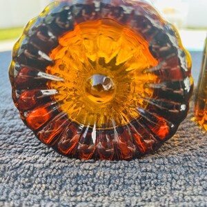 May include: Close-up of a decorative glass object with a flower-like design. The glass is amber and orange, with a dark brown rim. The object is sitting on a gray, textured surface.