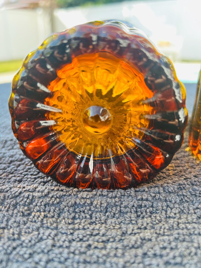 May include: Close-up of a decorative glass object with a flower-like design. The glass is amber and orange, with a dark brown rim. The object is sitting on a gray, textured surface.