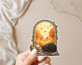 May include: A sticker featuring a cozy autumn scene. A black cat sleeps on books in front of a window overlooking a fall landscape with orange and yellow trees. A cup of tea and flowers are also present.