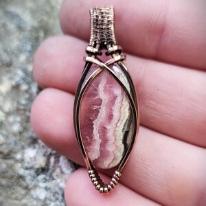 May include: A handcrafted pendant featuring a pink and white stone encased in copper wire. The wire is intricately woven at the top and bottom, with a decorative pattern. The stone has a layered appearance, with shades of pink, white, and gray.