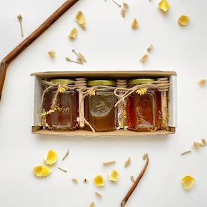 May include: Four small glass jars filled with honey, each with a wooden honey dipper and a gold ribbon tied around the lid. The jars are arranged in a row in a cardboard box.