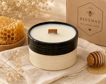 May include: A ceramic candle with a black and cream-colored design, featuring a wooden wick. The candle is surrounded by a honeycomb, a honey dipper, and a box labeled "100% Beeswax". The candle is on a neutral-colored surface.