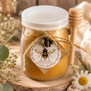 May include: A jar candle with a white lid, filled with yellow wax, and a wooden honey dipper. The jar is adorned with a twine bow and a round label featuring a bee illustration. The label reads "Pure Beeswax Candle".