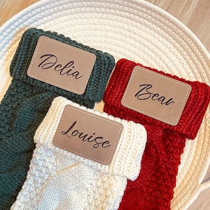 May include: Three knitted stockings with personalised leather name tags. The stockings are green, red and white. The names on the tags are Delia, Bear and Louise.