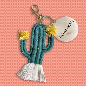 Pode incluir: Um chaveiro de cacto de fio verde com pompons amarelos e uma borla branca. O chaveiro tem um anel dourado e uma etiqueta de madeira que diz "SAVANNAH".