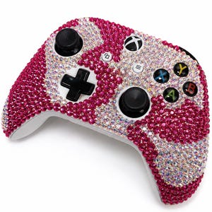 May include: A wireless video game controller fully encrusted with sparkling rhinestones in alternating patterns of vibrant hot pink and iridescent clear crystals. The device features black analog sticks, a black directional pad, and standard buttons.