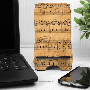 May include: A wooden phone stand featuring a vintage sheet music design. The stand is beige with black musical notes and rests on a white surface. A black smartphone and laptop are also visible.