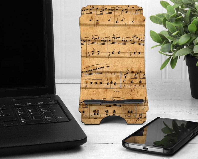 May include: A wooden phone stand featuring a vintage sheet music design. The stand is beige with black musical notes and rests on a white surface. A black smartphone and laptop are also visible.