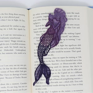 May include: Purple and blue embroidered mermaid bookmark. The mermaid is facing right and has a long flowing tail. The bookmark is made of lace and has a delicate design.