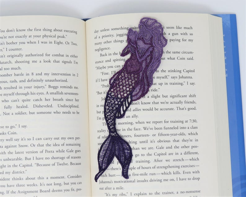 May include: Purple and blue embroidered mermaid bookmark. The mermaid is facing right and has a long flowing tail. The bookmark is made of lace and has a delicate design.