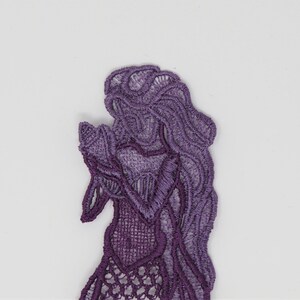 May include: Purple lace applique of a mermaid with long flowing hair. The mermaid is holding her hands together in front of her.
