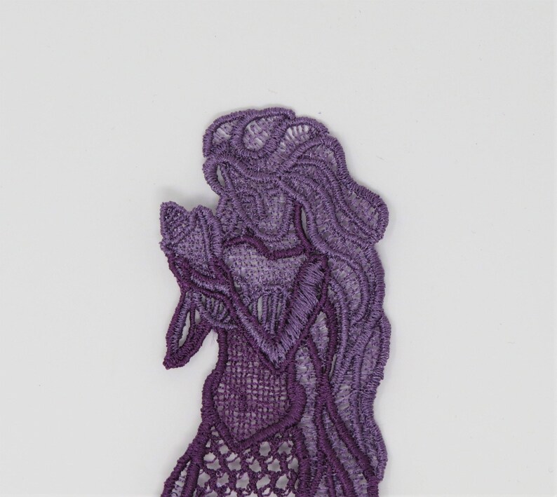 May include: Purple lace applique of a mermaid with long flowing hair. The mermaid is holding her hands together in front of her.