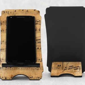 May include: A wooden phone stand with a vintage musical score design. The stand holds a black smartphone. The back of the stand is black. The stand is designed to hold a phone upright.