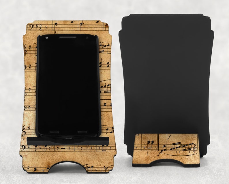 May include: A wooden phone stand with a vintage musical score design. The stand holds a black smartphone. The back of the stand is black. The stand is designed to hold a phone upright.
