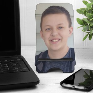 May include: A black phone stand with a photo of a young person with brown hair and a blue shirt. The stand is on a white surface.