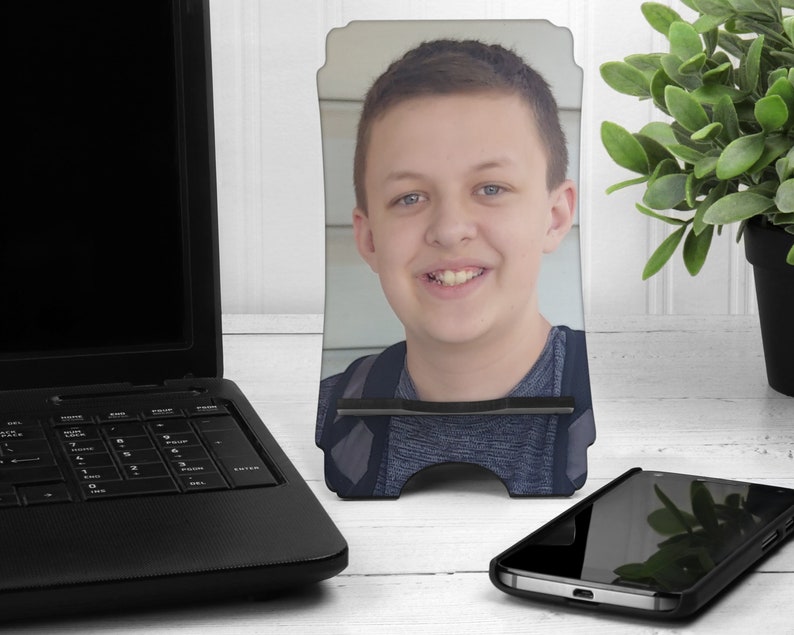 May include: A black phone stand with a photo of a young person with brown hair and a blue shirt. The stand is on a white surface.