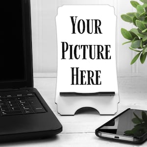 May include: White phone stand with the text "YOUR PICTURE HERE" on it. The stand is on a white wooden surface with a laptop and a smartphone next to it.